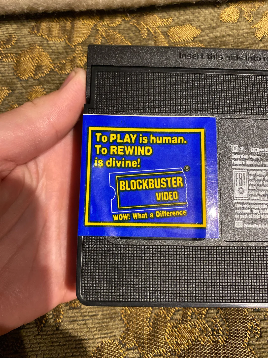 A hand holds a VHS tape with a blue and yellow Blockbuster Video sticker that says, “To PLAY is human. To REWIND is divine! WOW! What a Difference.”