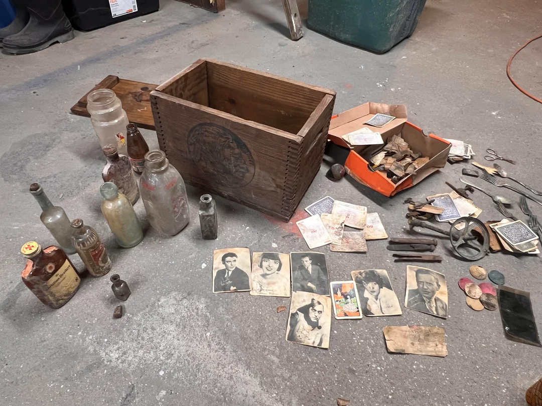 A wooden crate sits on a concrete floor surrounded by old bottles, jars, black-and-white photos, playing cards, keys, buttons, and other assorted vintage items spread out nearby.