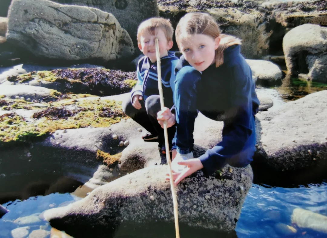 Two children, a girl and a boy, kneel on rocks near water. The girl holds a long stick over the water. Both wear blue jackets. The scene appears to be outdoors on a sunny day, possibly by the sea.