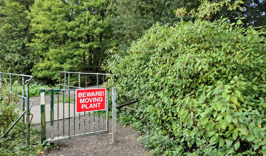 A metal gate with a red sign reading “BEWARE! MOVING PLANT” stands beside dense green bushes along a pathway in a wooded area.