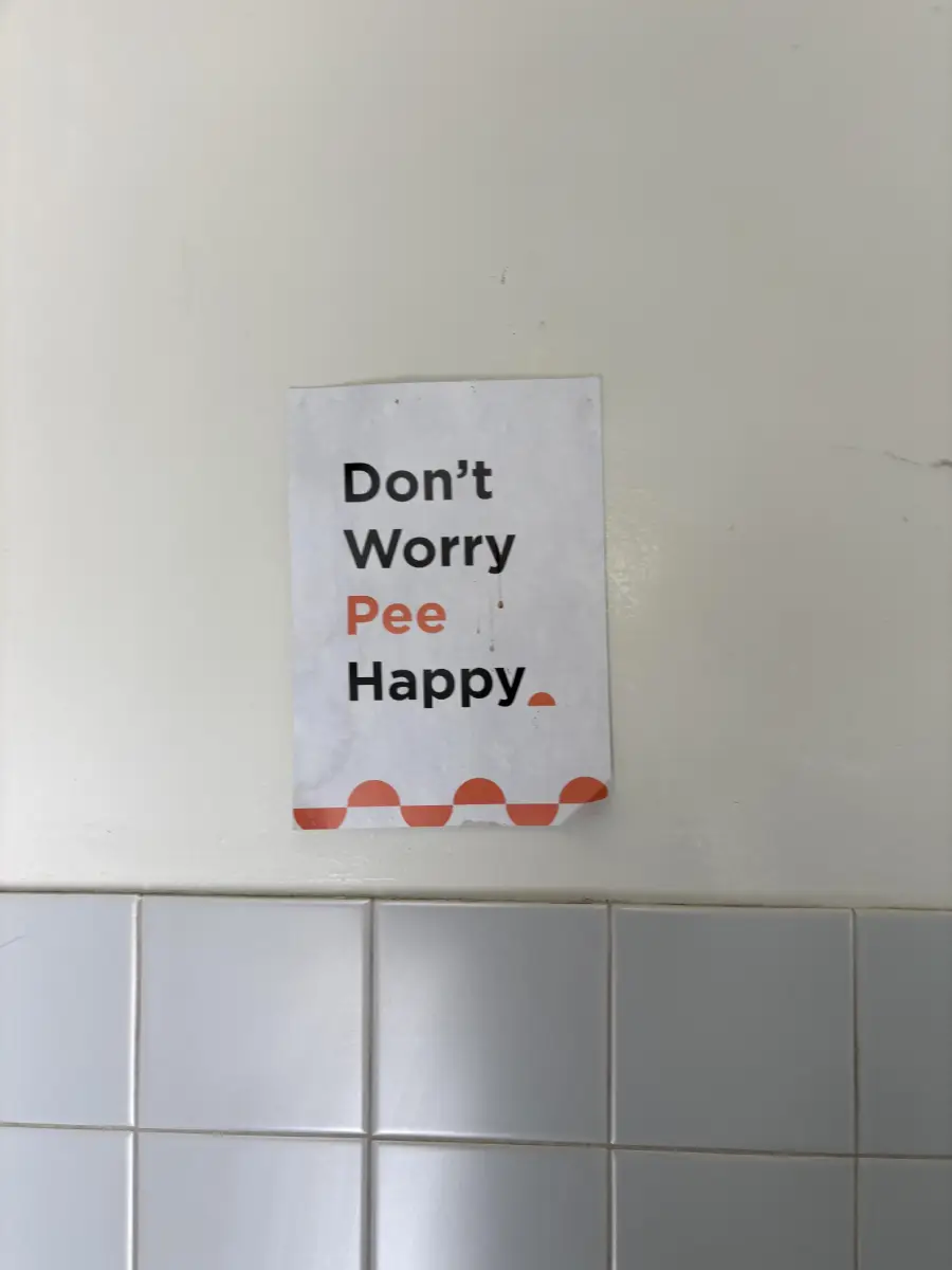 A sign on a tiled restroom wall reads, “Don’t Worry Pee Happy,” with the word “Pee” in orange and the rest in black.