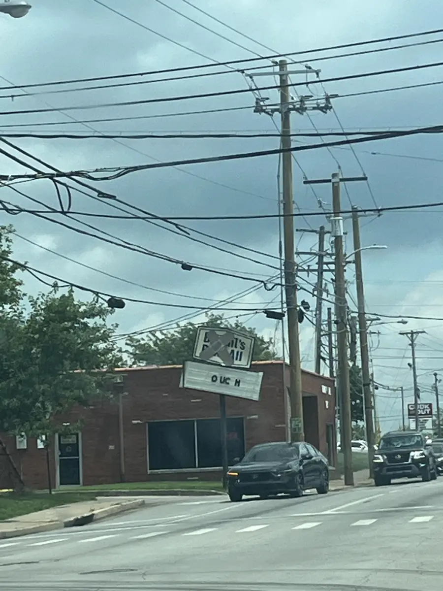 A brick building with a sign reading “DUCK” stands at a street corner. Power lines crisscross overhead, and cars are driving along the road under a cloudy sky.