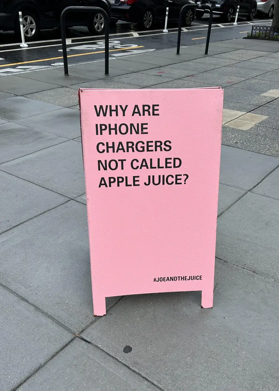 A pink sidewalk sign reads, “WHY ARE IPHONE CHARGERS NOT CALLED APPLE JUICE?” with the hashtag #JOEANDTHEJUICE at the bottom. The sign is on a city sidewalk near the street.