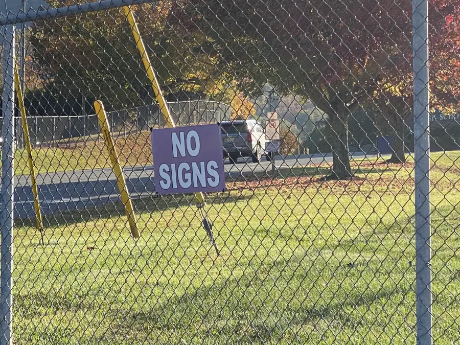 A chain-link fence with a sign that ironically reads “NO SIGNS” placed on a grassy field near a road, with trees and a white vehicle in the background.