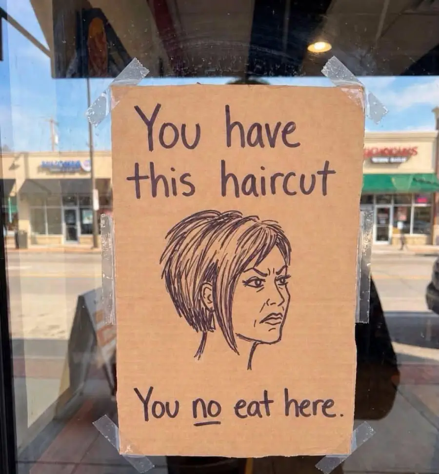 A cardboard sign taped to a window shows a drawing of a woman with an angled bob haircut and reads: “You have this haircut, you no eat here.” Stores are visible in the background through the glass.