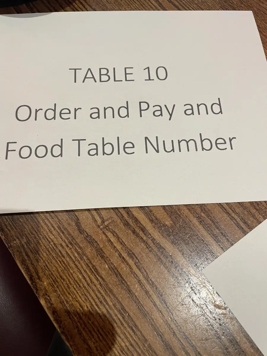 A paper sign on a wooden table reads: “TABLE 10 Order and Pay and Food Table Number.”