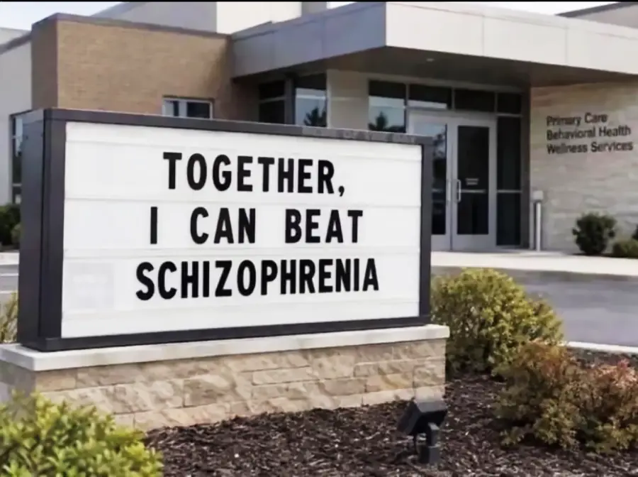 A sign outside a healthcare building reads, “TOGETHER, I CAN BEAT SCHIZOPHRENIA.” Bushes are planted in front of the sign. The entrance and signage of the building are visible in the background.