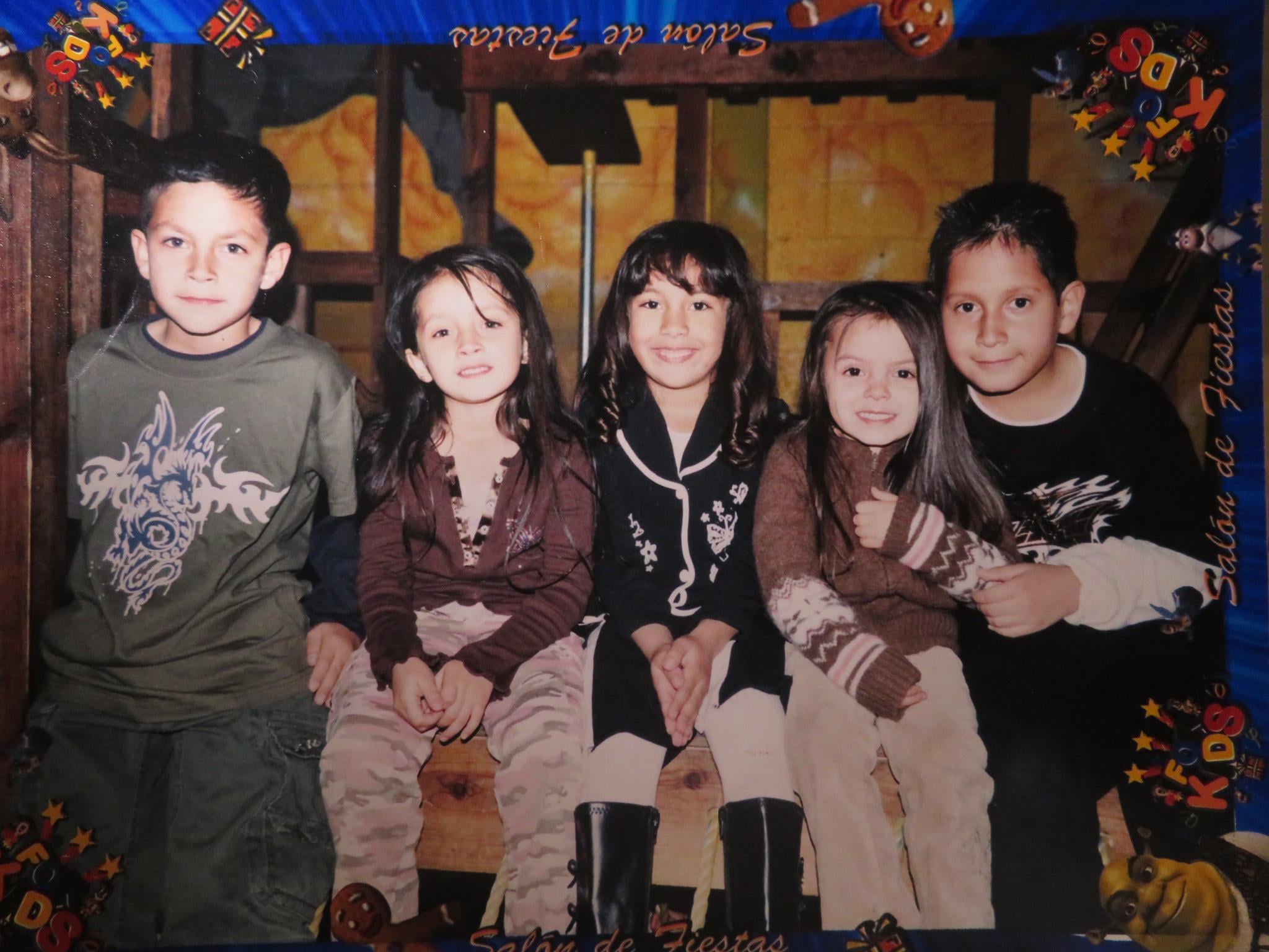 Five children sit closely together on a wooden bench, smiling at the camera. The photo has a colorful party-themed border with cartoon decorations and Spanish text, suggesting a festive or birthday celebration.