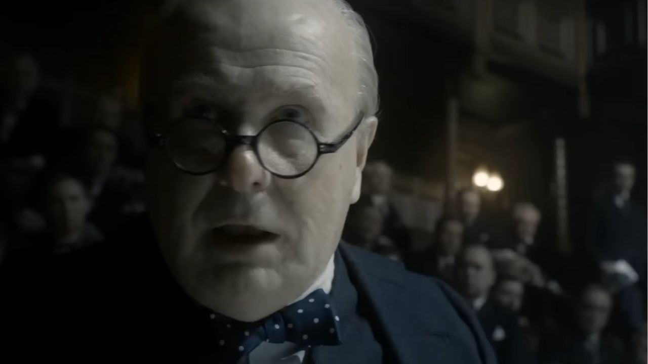 A man with round glasses, a bow tie, and a serious expression speaks indoors, with several people sitting in the dimly lit background.