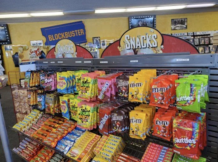 A display of colorful candy bags and chocolate bars in front of a Blockbuster sign, with “Snacks” labels above the shelves and DVDs on racks in the background.