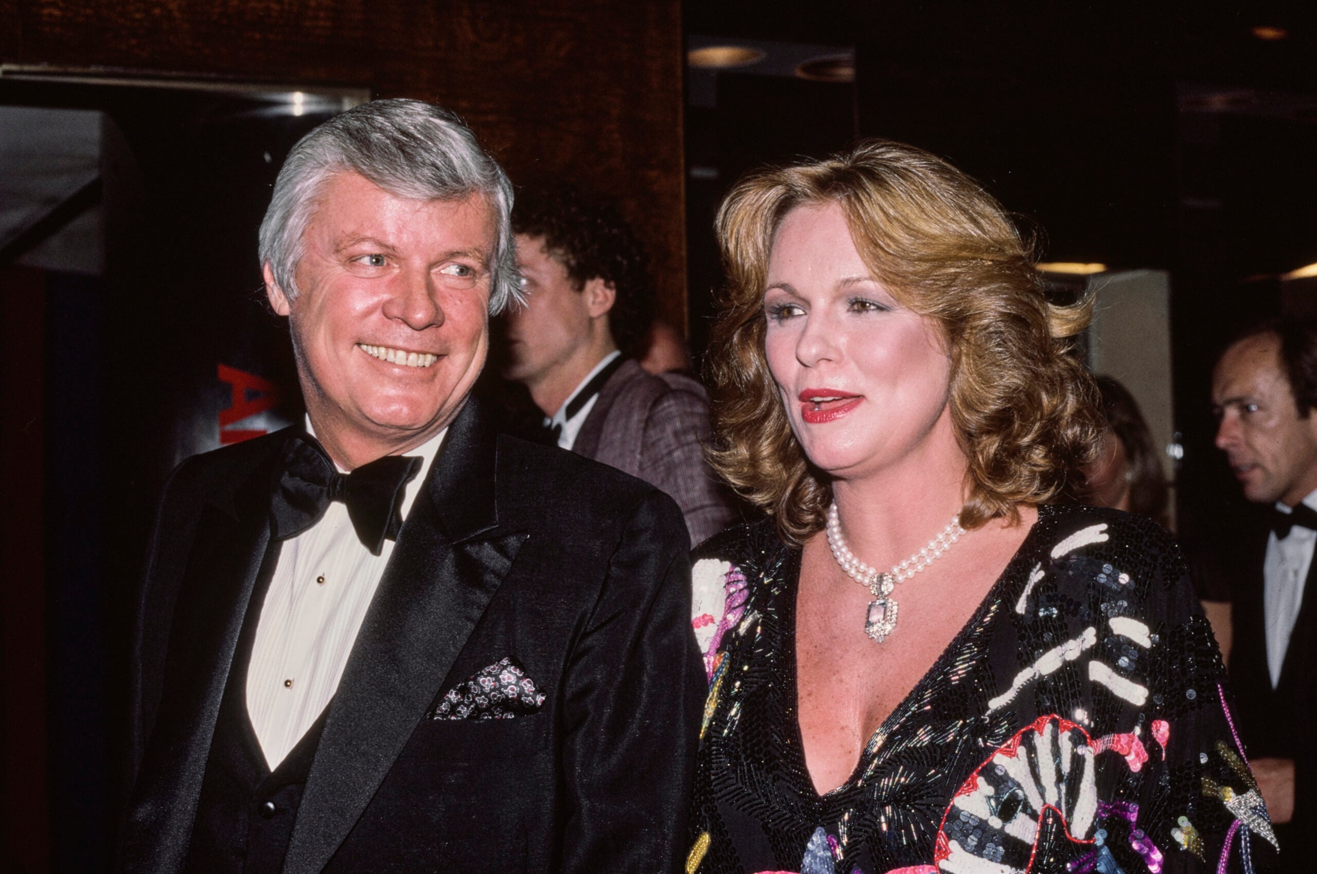 A man in a black tuxedo and a woman in a colorful dress with a pearl necklace smile at a formal event. Other guests in formal attire are in the background.