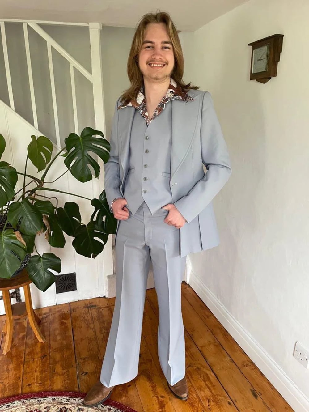 A smiling person with long hair wears a light blue three-piece suit and brown shoes, standing indoors on a wooden floor near a staircase and a leafy green plant.