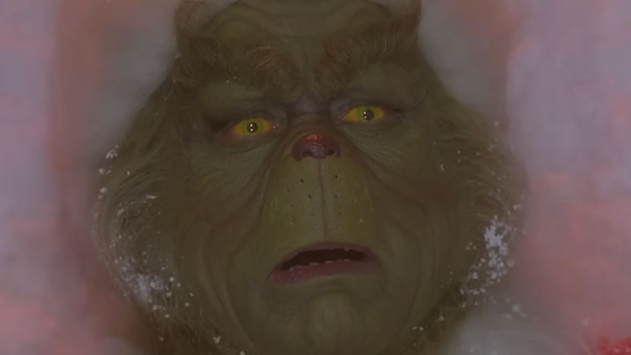 A close-up of the Grinch’s green, furry face with yellow eyes, furrowed brows, and a red nose, surrounded by white fur trim and a dusting of snow.