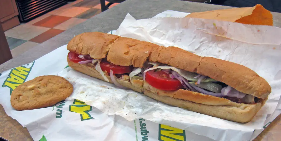 A Subway sandwich with tomatoes, onions, lettuce, and pickles sits on branded paper next to a chocolate chip cookie on a table.