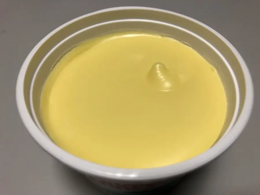 An opened tub of smooth yellow margarine with a small swirl or bump on its surface, sitting on a gray background.