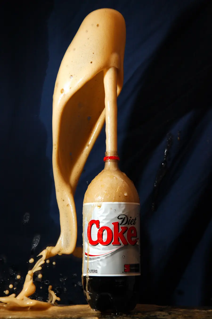 A 2-liter bottle of Diet Coke erupts with foam, shooting a geyser of fizz high into the air against a dark background.