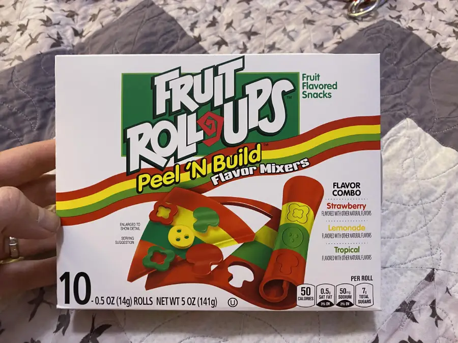 A hand holds a box of Fruit Roll-Ups Peel ’N Build Flavor Mixers, showing colorful fruit snack shapes and flavors: strawberry, lemonade, and tropical. The box contains 10 rolls.