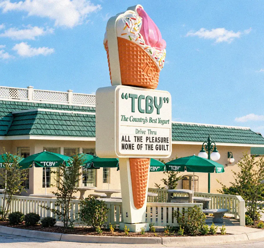 A large ice cream cone sign outside a TCBY yogurt shop reads: “TCBY, The Country’s Best Yogurt. Drive Thru. All the pleasure, none of the guilt.” Green awnings and outdoor umbrellas surround the building.