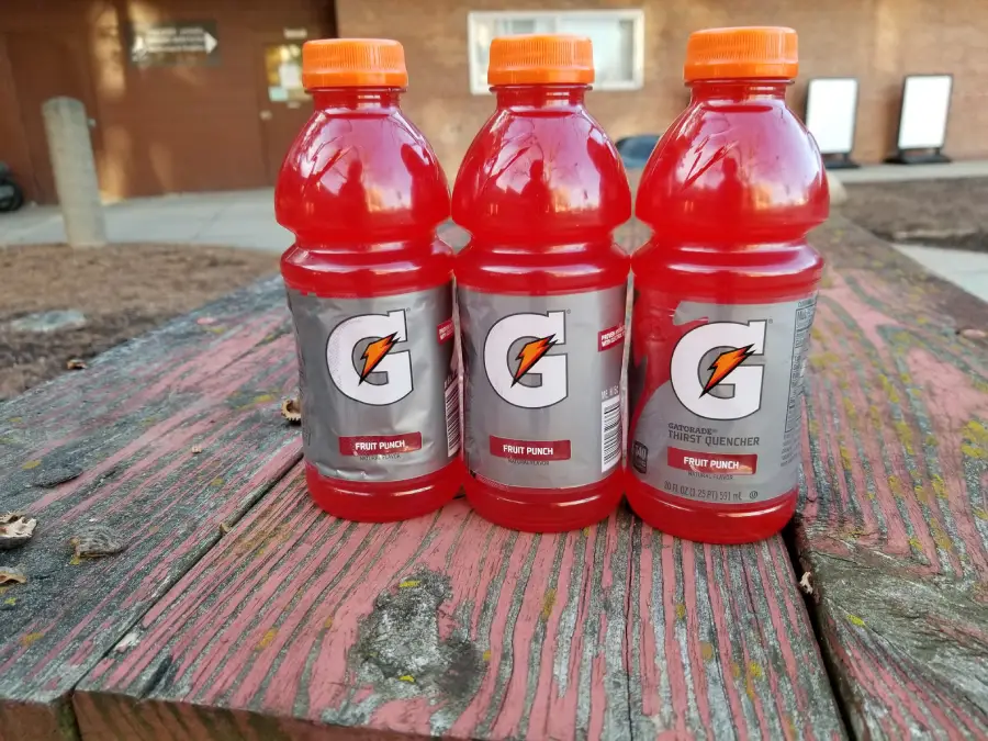 Three bottles of red Gatorade Fruit Punch are placed side by side on an old, weathered wooden picnic table outdoors, with a building and signs visible in the background.