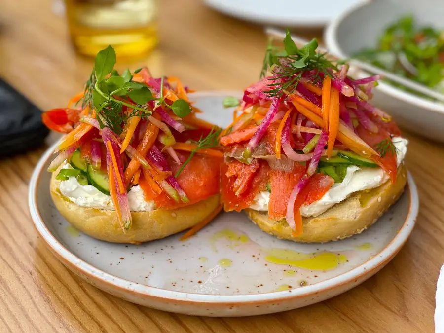 Two bagel halves topped with cream cheese, smoked salmon, sliced red onions, shredded carrots, radishes, and fresh herbs, served on a white plate with a drizzle of olive oil.