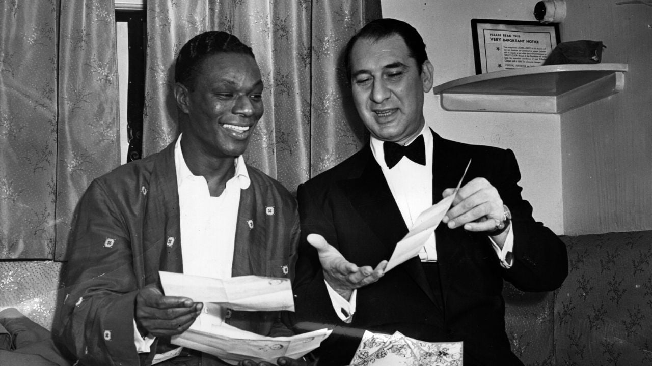 Two men sit on a couch smiling and looking at papers together; one wears a dark robe, the other is in a tuxedo. There are curtains and a framed document in the background.