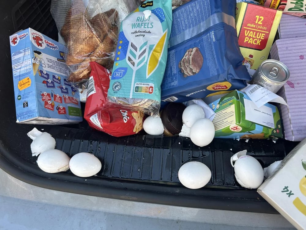 Several groceries, including snacks, canned food, and bread, are scattered in a car trunk. Six eggs, some of them broken, are lying among the items, creating a mess.