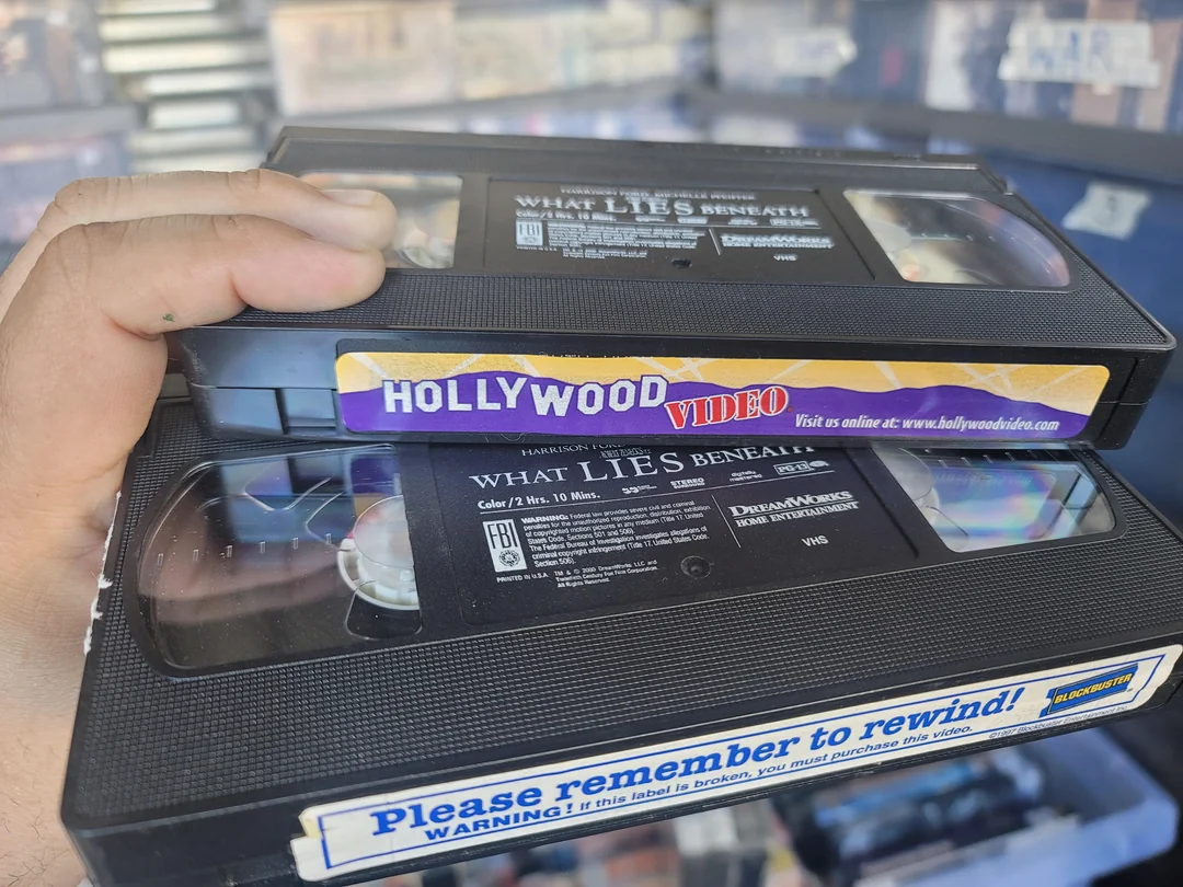 A hand holds two VHS tapes from Hollywood Video. The tapes have labels for the movie “What Lies Beneath” and a sticker reminding to rewind. Shelves of other VHS tapes are blurred in the background.