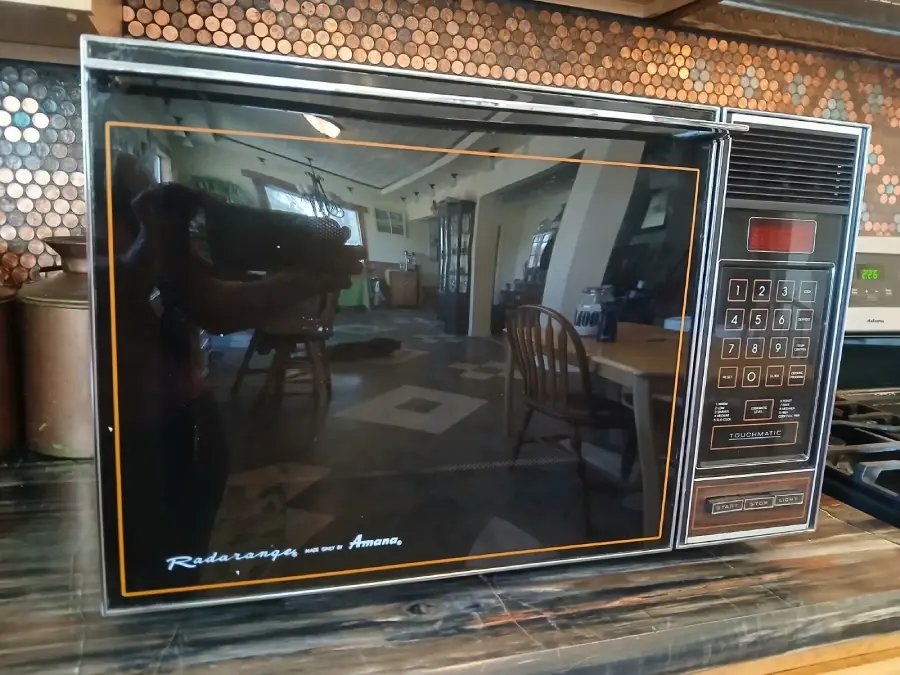 Vintage Amana Radarange microwave oven with a black glass door, keypad controls, and a digital display, sitting on a kitchen counter with reflections of chairs and a table visible in the door.