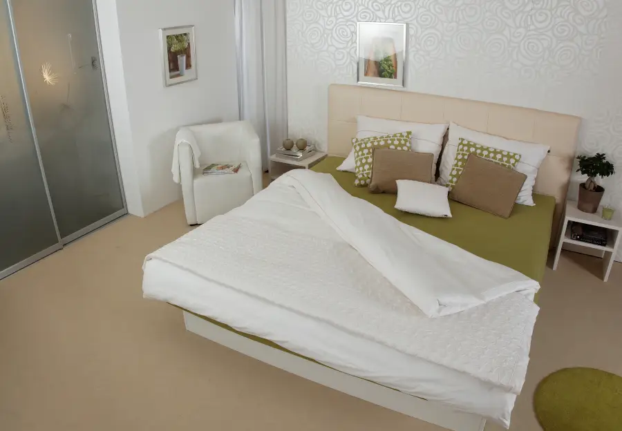A modern bedroom with a green and white bed, multiple throw pillows, a white armchair with a book, bedside tables, a potted plant, sliding closet doors, and framed artwork on patterned white walls.