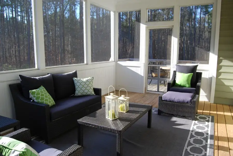 Screened-in porch with dark wicker furniture, green and patterned pillows, a rug, a coffee table with two lanterns, and a wooded view outside. Sunlight streams through the large windows.