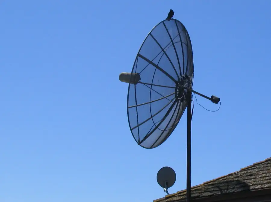 A large satellite dish with a small bird perched on its rim, set against a clear blue sky. A smaller satellite dish and part of a roof are visible at the bottom right of the image.