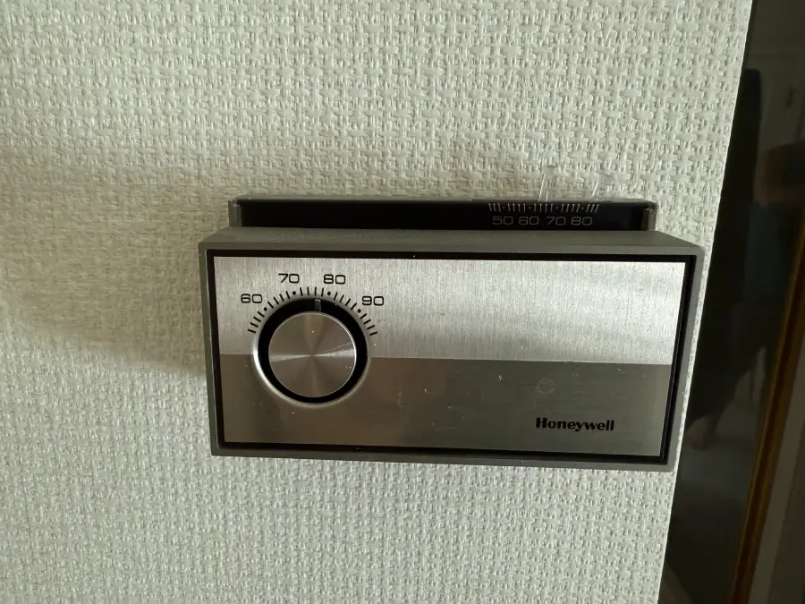 A Honeywell wall thermostat with a round dial set to about 77°F, mounted on a textured white wall.