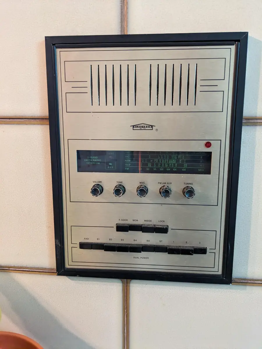 A vintage wall-mounted intercom and radio system with dials, buttons, and a speaker grille, set in a tiled wall.