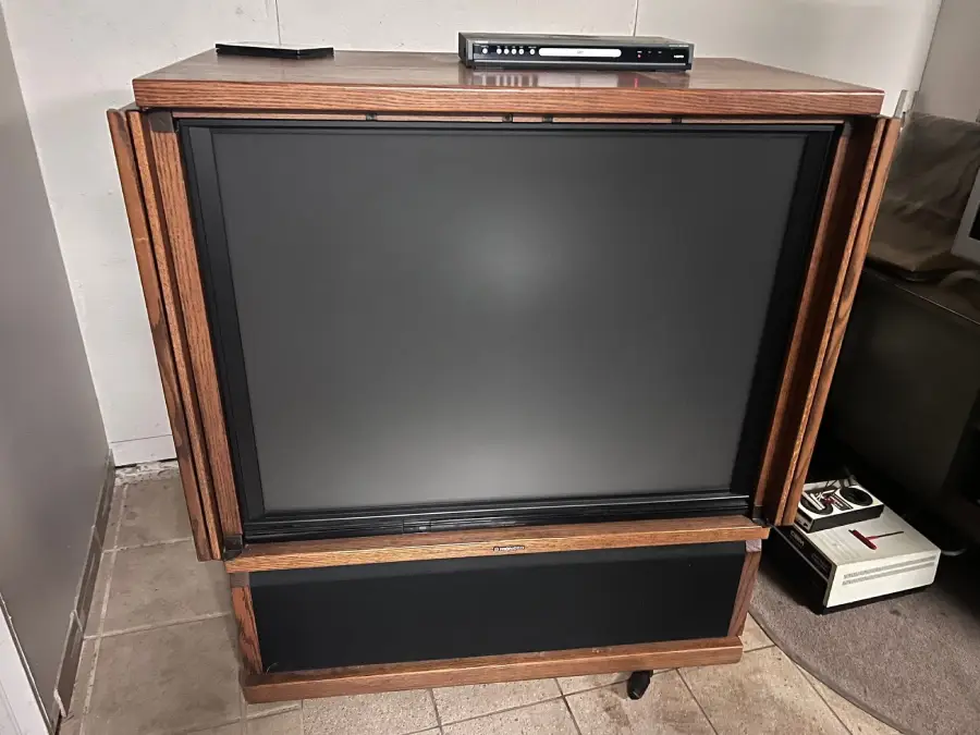 A vintage large wooden console television with a black screen, a remote on top, and an electronic device. A VCR sits on the floor nearby. The scene is in a tiled room.