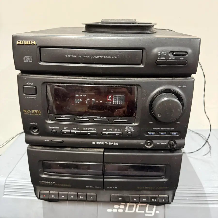 A black Aiwa stereo system with a CD player on top, a central control panel, volume knob, display screen, buttons, and two cassette tape decks at the bottom. The unit is on a reflective surface.