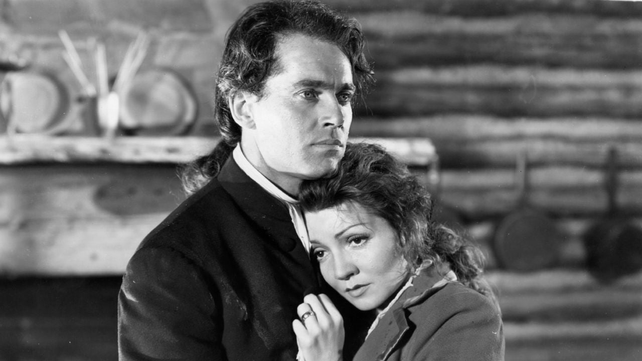 A man and woman, both dressed in old-fashioned clothing, stand close together inside a rustic cabin. The woman rests her head on the man’s chest, looking concerned, while the man stares ahead seriously.