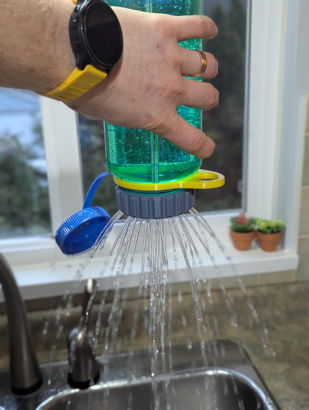 A hand holds an upside-down water bottle with a round sprinkler cap, creating multiple streams of water over a kitchen sink. A window, faucet, and potted plants are visible in the background.