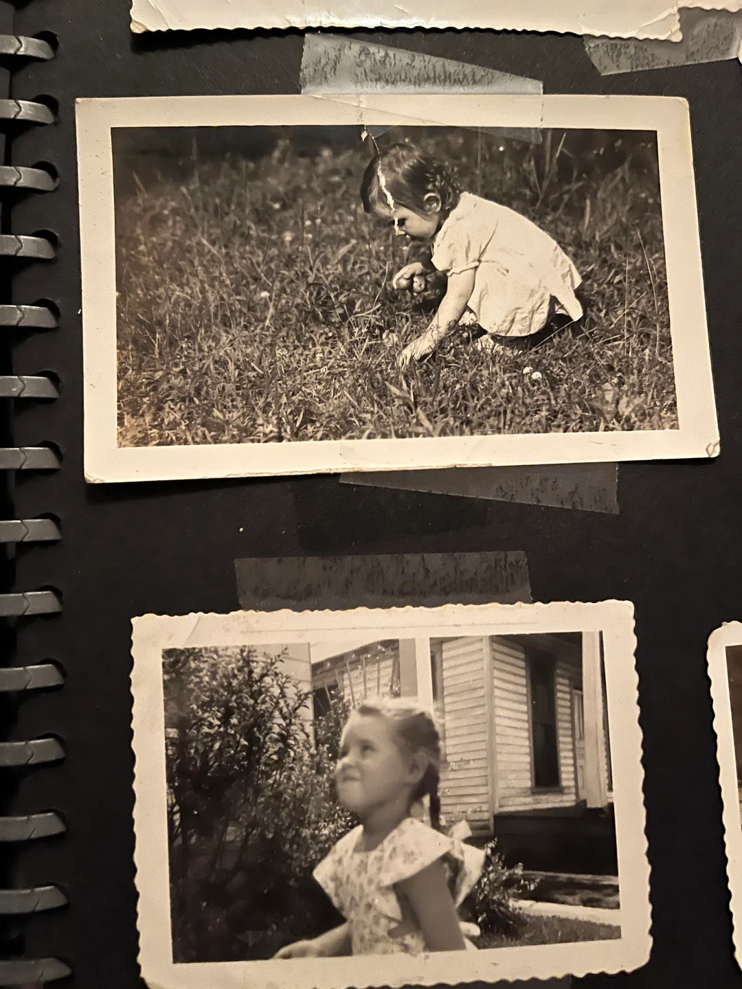 Two black-and-white photos in a scrapbook: a young child sits on grass in the top photo; below, a girl in a light-colored dress sits outside near a house, both images bordered with white edges.