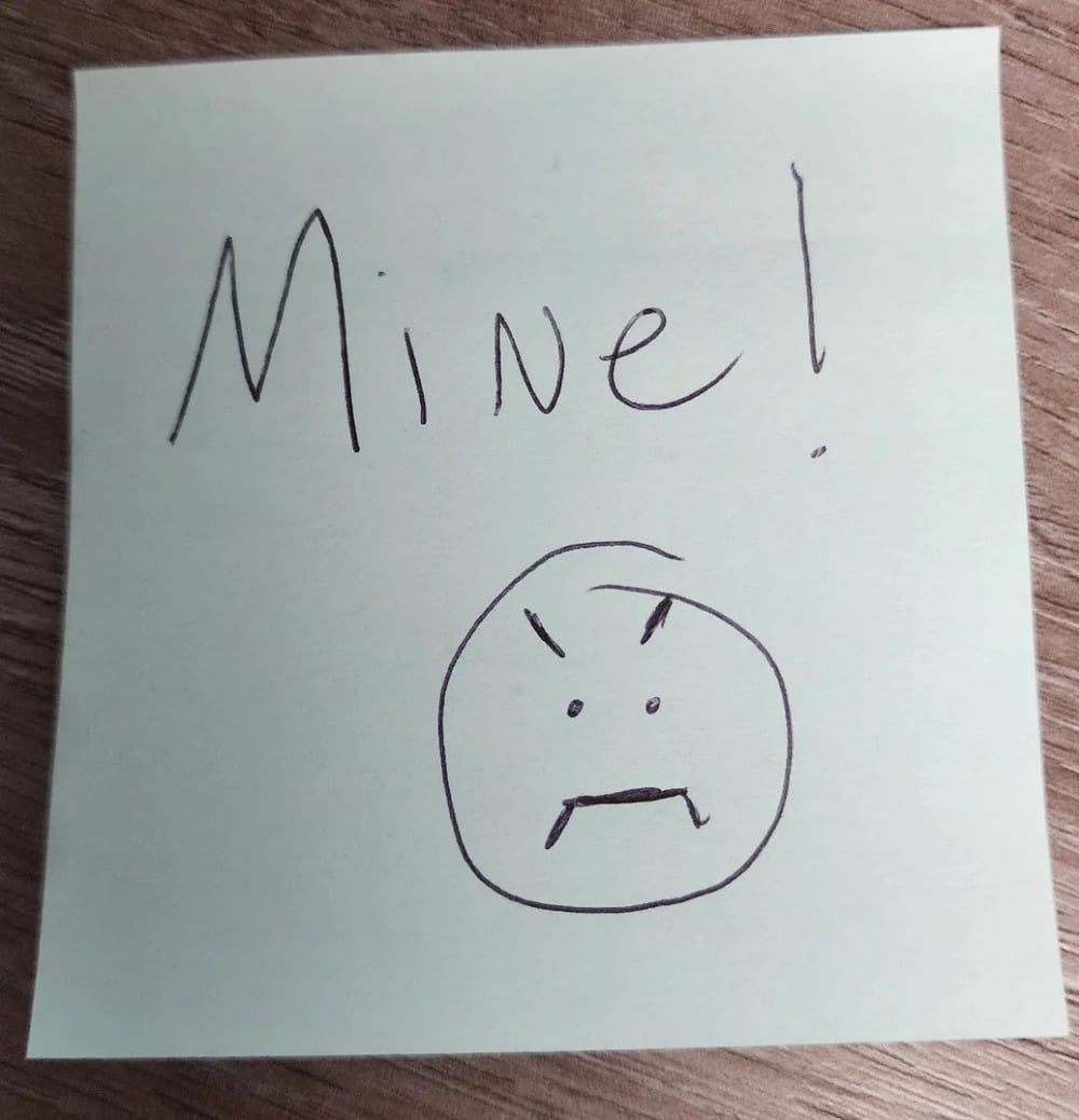 A sticky note reads “Mine!” above a simple drawing of an angry face with furrowed brows and a frown, placed on a wooden surface.