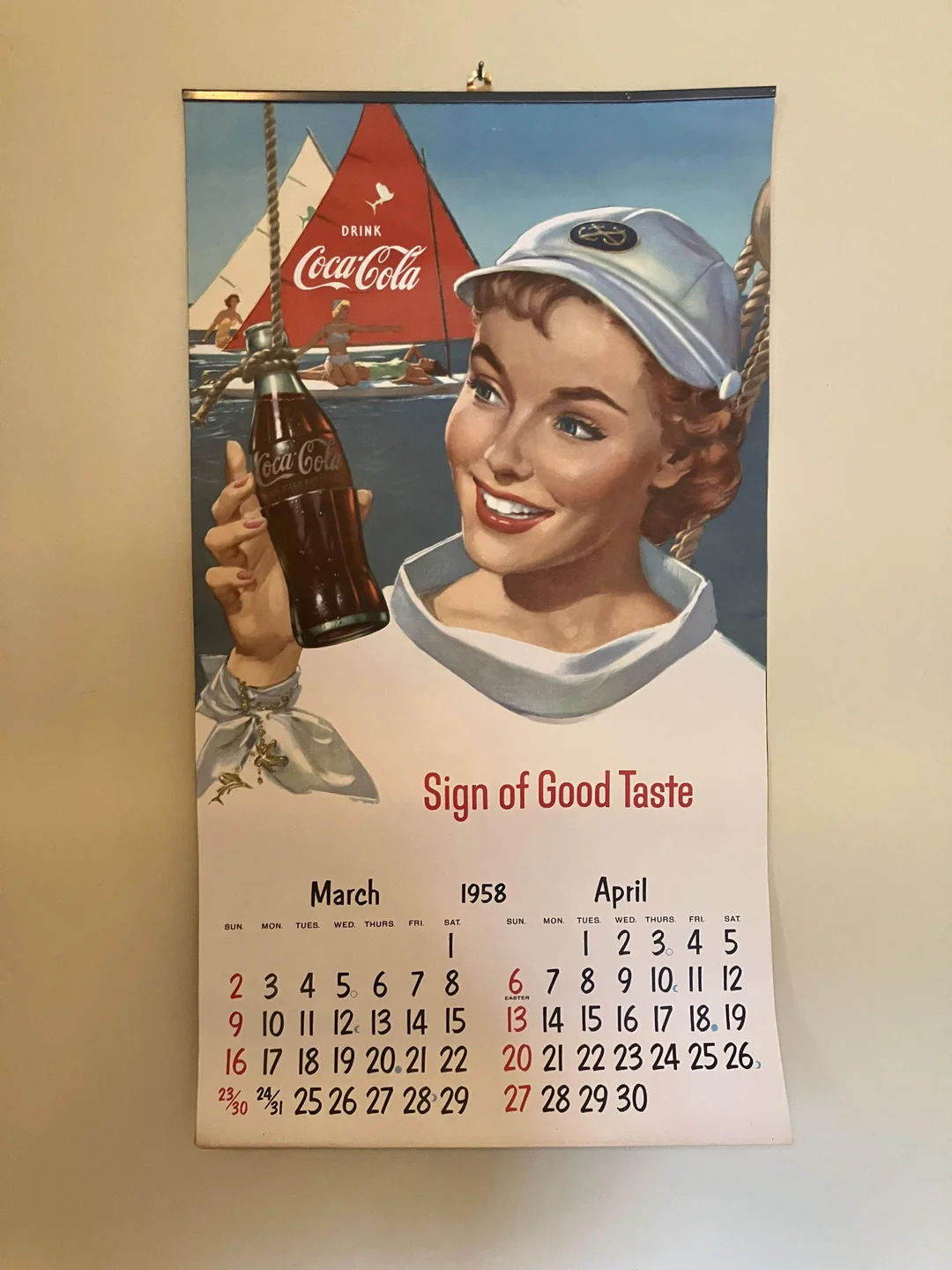 A vintage 1958 Coca-Cola calendar shows a smiling woman in a sailor outfit holding a Coke bottle, with a sailboat and “Drink Coca-Cola” sign in the background. March and April dates are visible below.