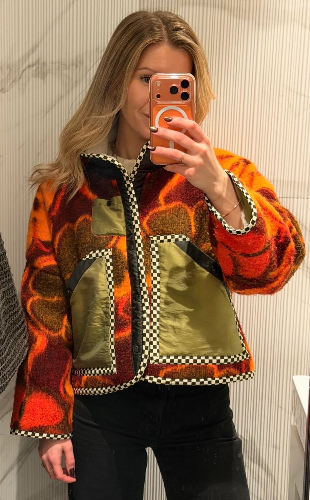 A person takes a mirror selfie wearing a colorful jacket with orange, red, and yellow patterns, large floral designs, and shiny green pockets. The background shows a white, striped wall.