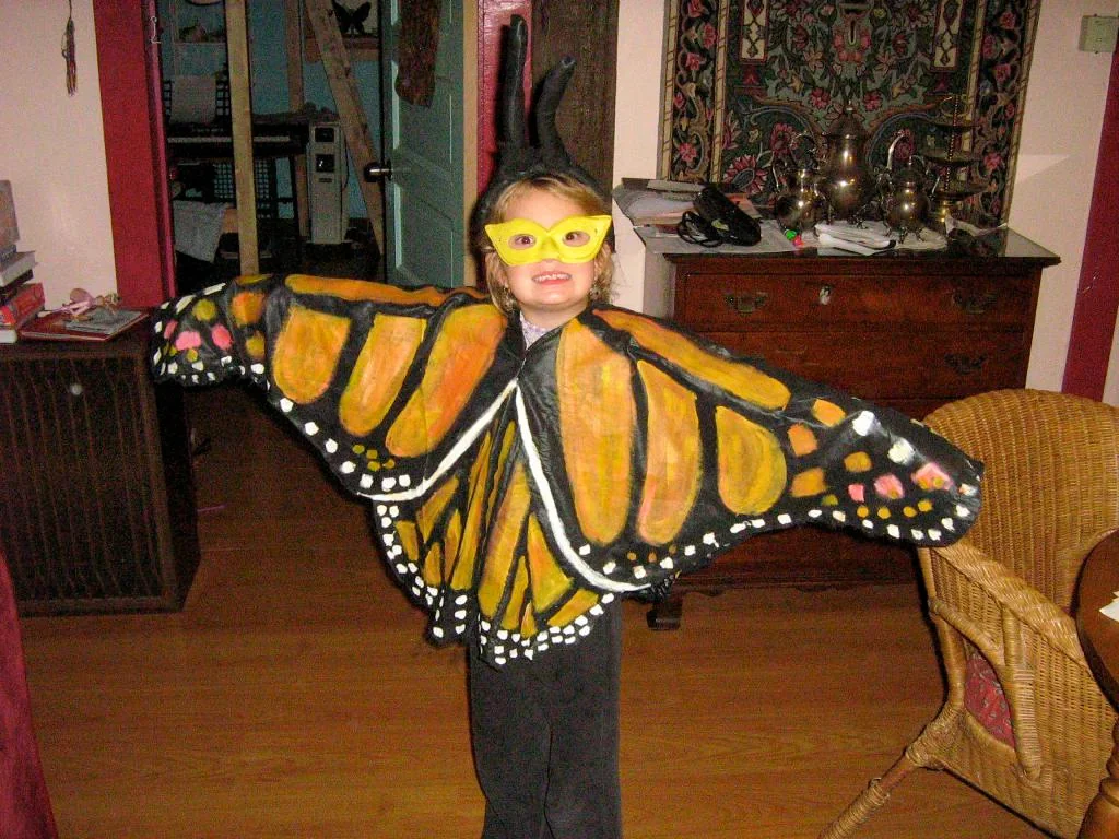 A child wearing a monarch butterfly costume with bright yellow and orange wings, black clothing, and yellow mask, standing indoors with arms spread wide.
