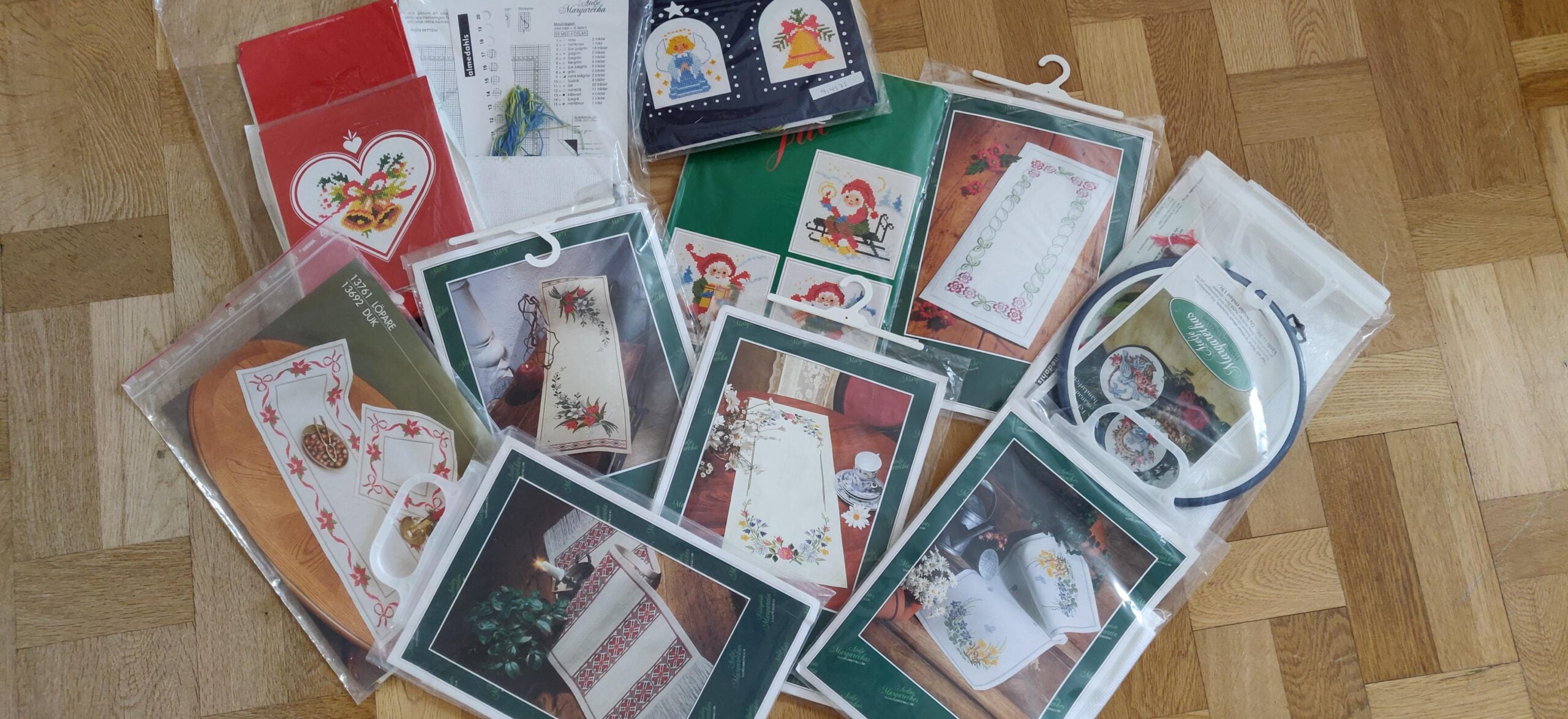 Assorted embroidery kits and patterns, many with Christmas or festive designs, are spread out on a wooden parquet floor. Each kit is in clear plastic packaging and some feature images of the finished embroidered items.
