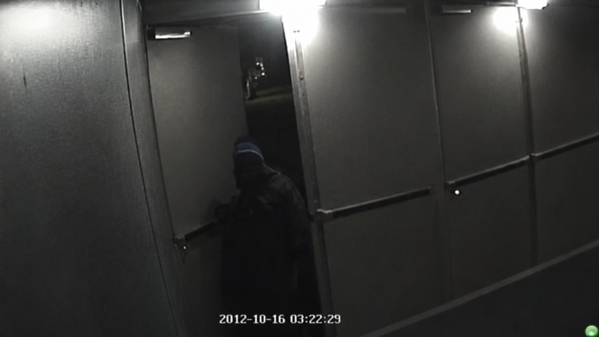 A person in dark clothing and a blue hat enters a building at night through double doors. The timestamp reads “2012-10-16 03:22:29.” The scene is dimly lit by overhead lights.