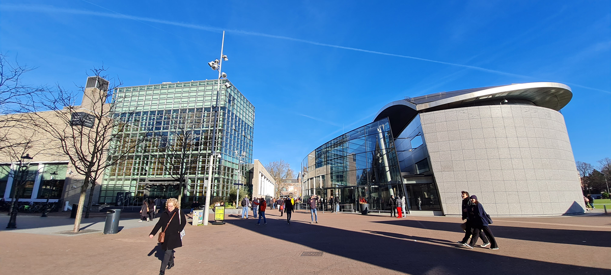 People walk between two modern glass buildings under a clear blue sky, with trees and more buildings visible in the background.