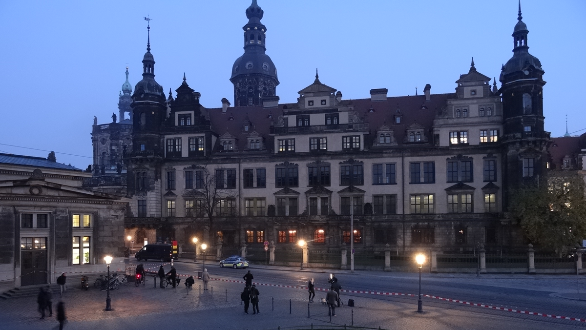 People walk near a historic European building at dusk. The building has towers and many windows. Street lamps are lit, and police vehicles are parked along the nearly empty street. The sky is a hazy blue.