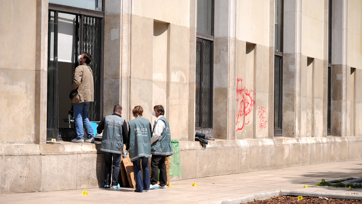 Four people in police vests examine a building’s exterior near tall windows with evidence markers on the ground. One person stands on a ledge looking up, while red graffiti is visible on the wall.