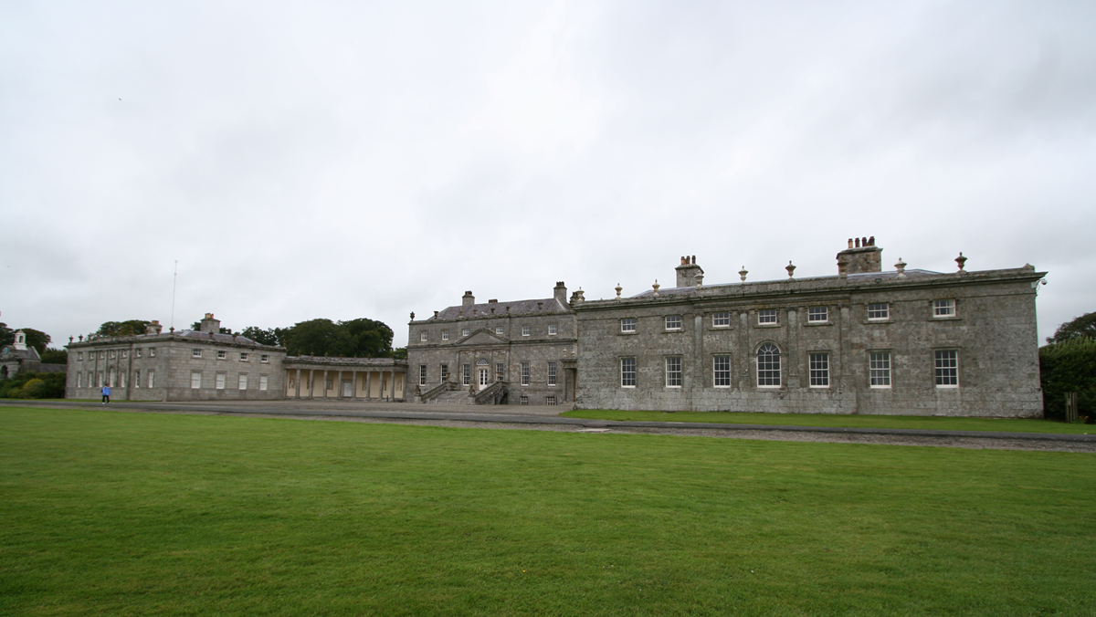Large, historic stone building with many windows, chimneys, and columns, set against a cloudy sky. A wide, green lawn stretches across the foreground.