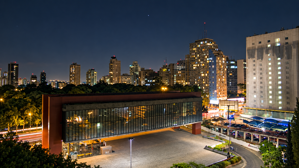 Night view of São Paulo’s MASP museum with its distinct glass structure, surrounded by tall, illuminated buildings and trees, under a clear sky. Busy city lights and traffic are visible in the background.