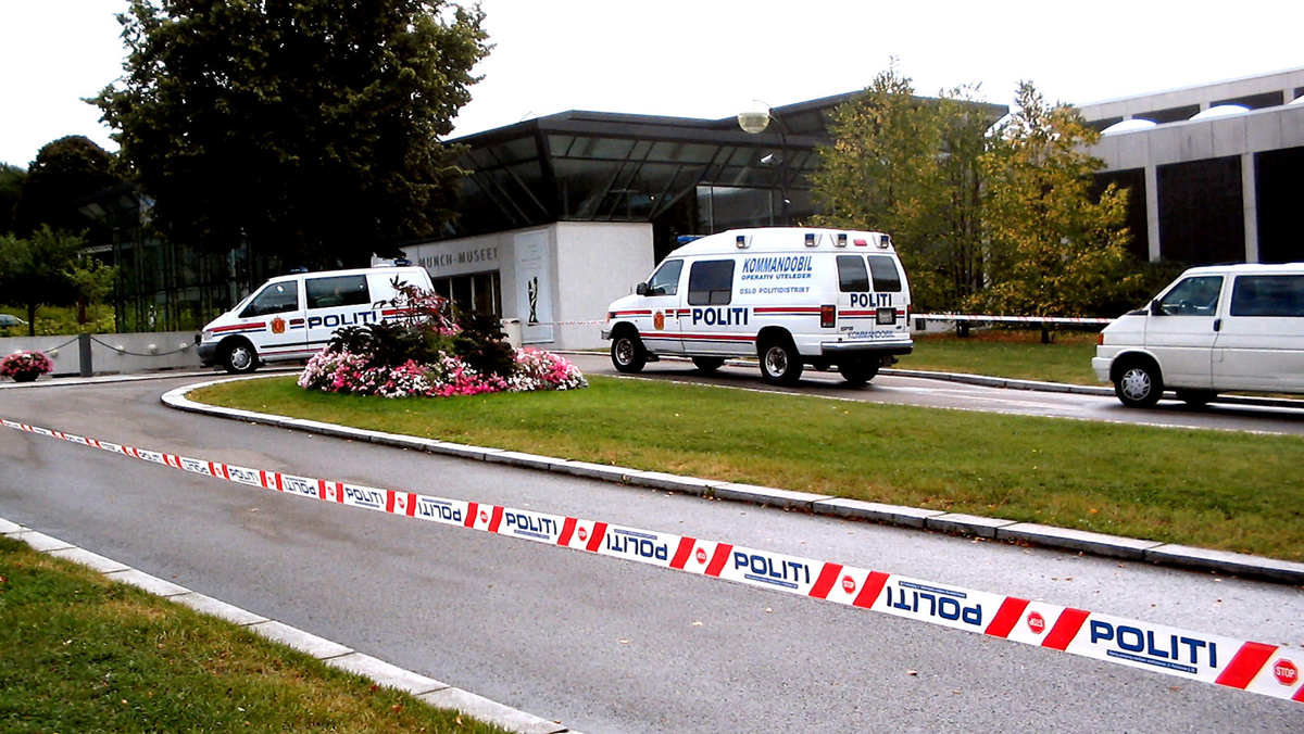 Two police vehicles and a white van are parked near a modern building, behind police tape labeled “POLITI.” There are flower beds and trees in the grassy area by the entrance.